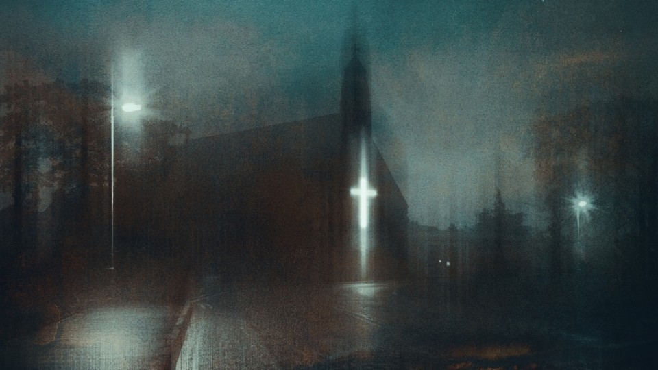 A color photograph of a church with a bright cross light seen from across the street on a gloomy blue night.