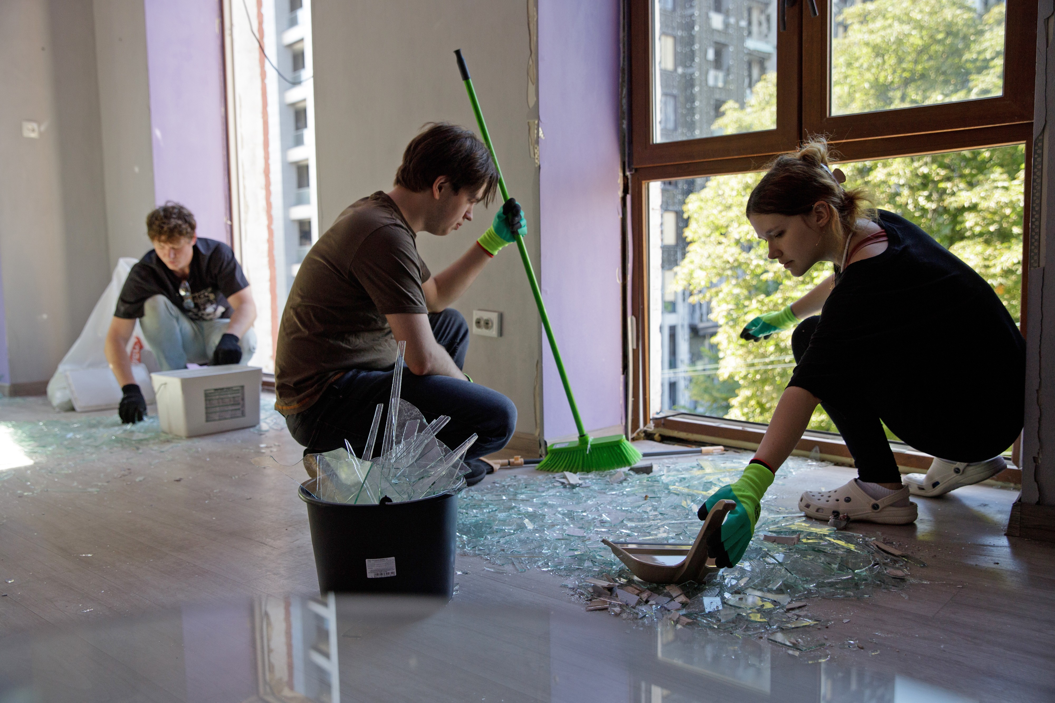 Several people collect glass shards beside broken windows in a damaged building.