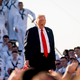 President Donald Trump is shown speaking, with his mouth open, surrounded by uniformed members of the U.S. Navy.