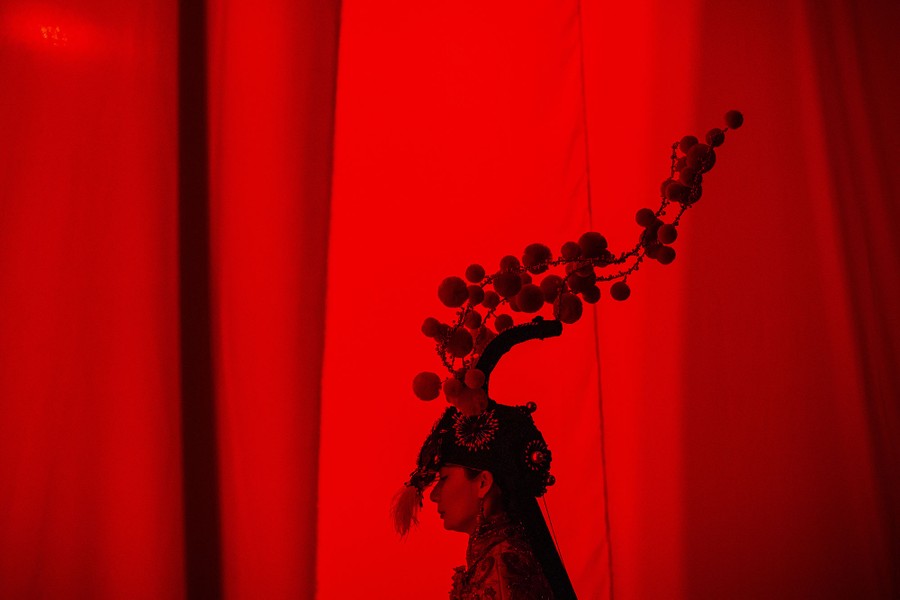 A model in a tall headdress stands behind a red curtain.