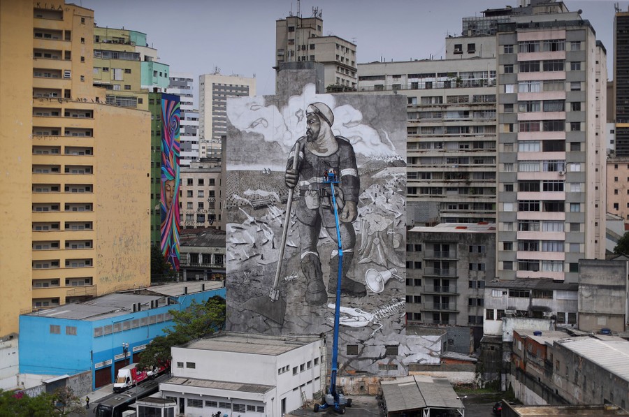 An artist on a tall crane works on a very tall mural on the side of a building in a city.