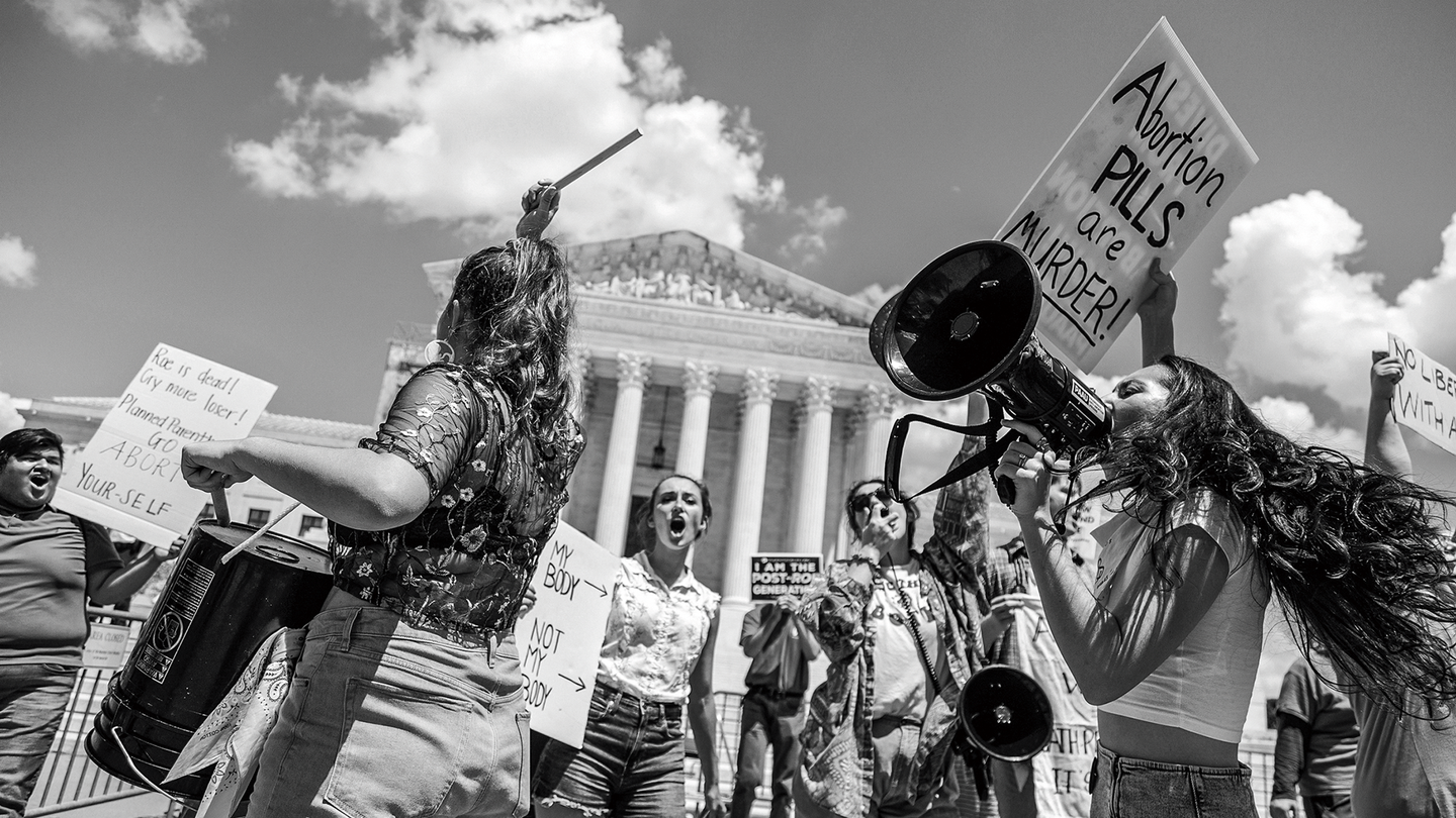 Black-and-white photo of anti-abortion protesters drumming and shouting through bullhorns in front of the Supreme Court, holding signs, including one that reads "Abortion PILLS are MURDER!"