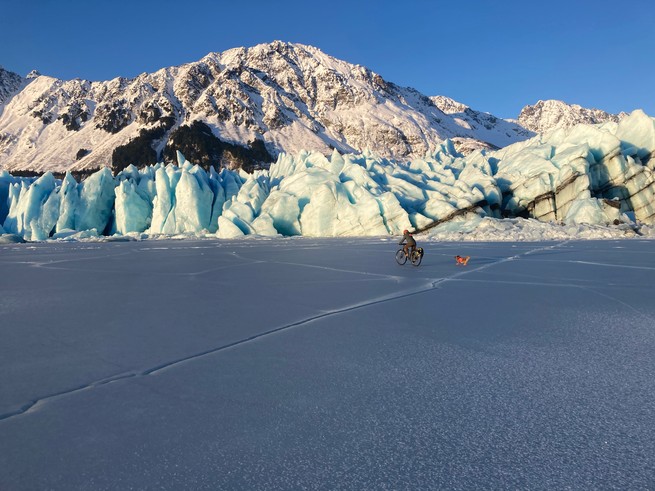 A woman rides her bike across an ice-locked lake, chased by a tiny dog