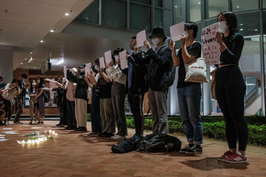 About a dozen protesters stand side by side near a building, holding up signs or blank pieces of paper.