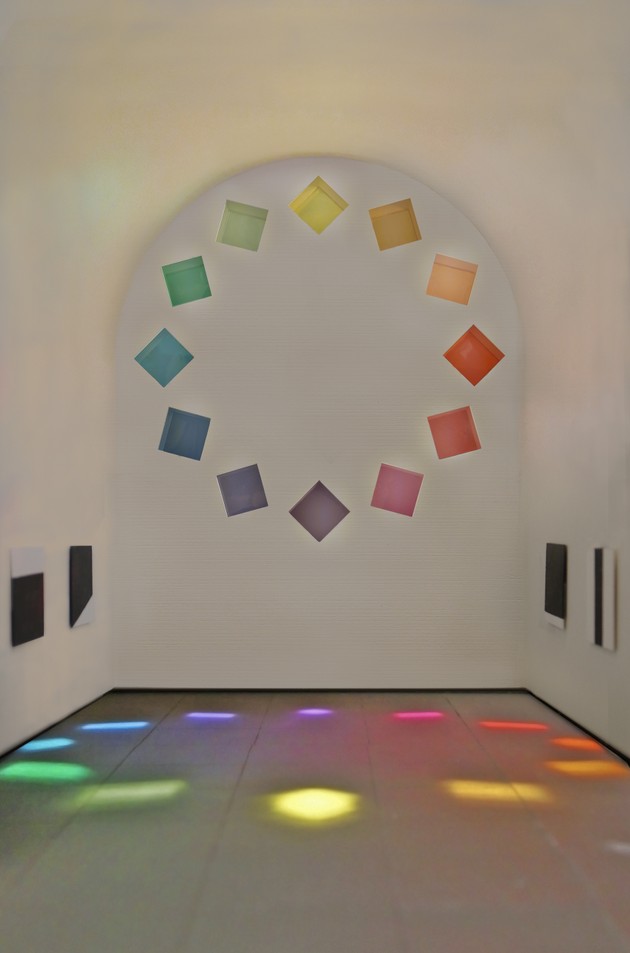 A white room with a rainbow of lights against the back wall. The colors of the lights reflect onto the ground 