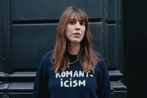 Alice Birch stands against a black wall with her hair blowing, wearing a blue sweater that reads "Romanticism."
