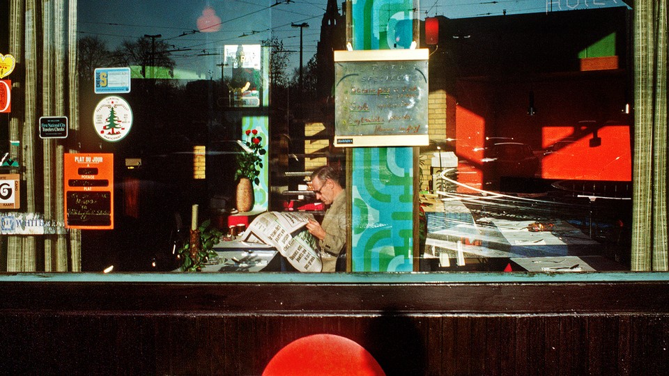 A man, photographed through a large window, reads a newspaper by himself in a restaurant.