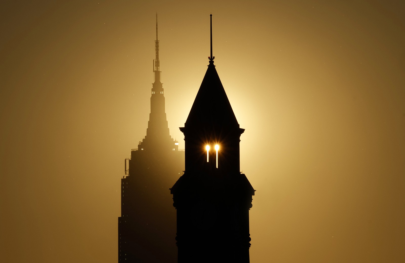 The sun rises behind the Empire State Building and another tower, silhouetting the buildings.