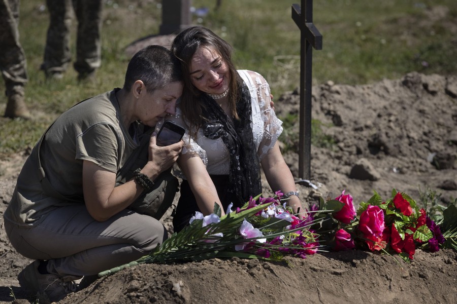 Two people kneel beside a flower-covered grave.
