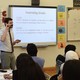 A teacher stands near a whiteboard that describes "Learning Goals" and another board with math problems; students sit at desks.