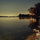 photo at night of lake shore with grassy bank, glowing horizon, and sky with bright stars