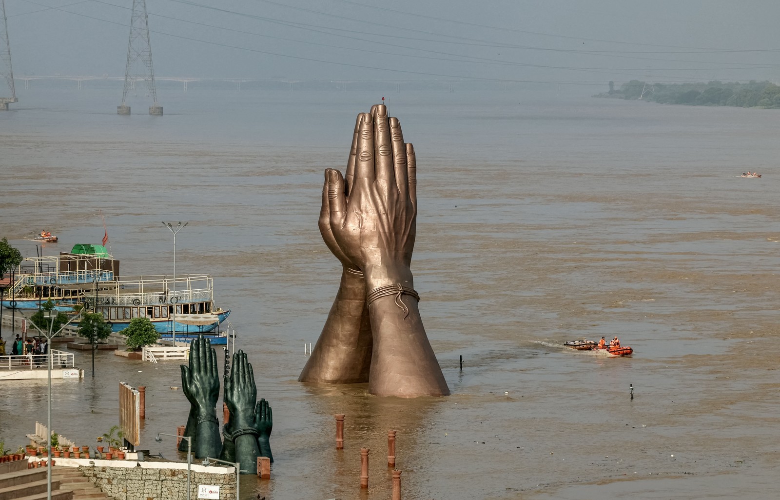 A large statue of hands placed together, seen along a flooded riverbank.