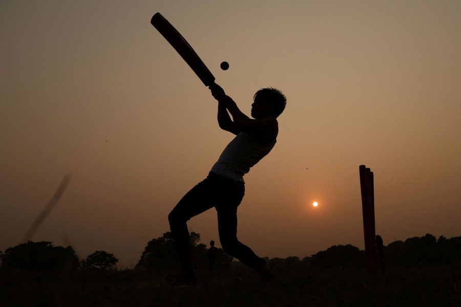 A person swings a cricket bat at a ball, silhouetted by the low sun.