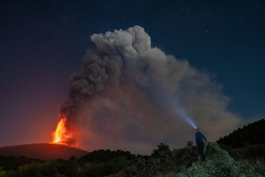 A person wearing a headlamp watches ash and lava erupting from Mount Etna in the distance.