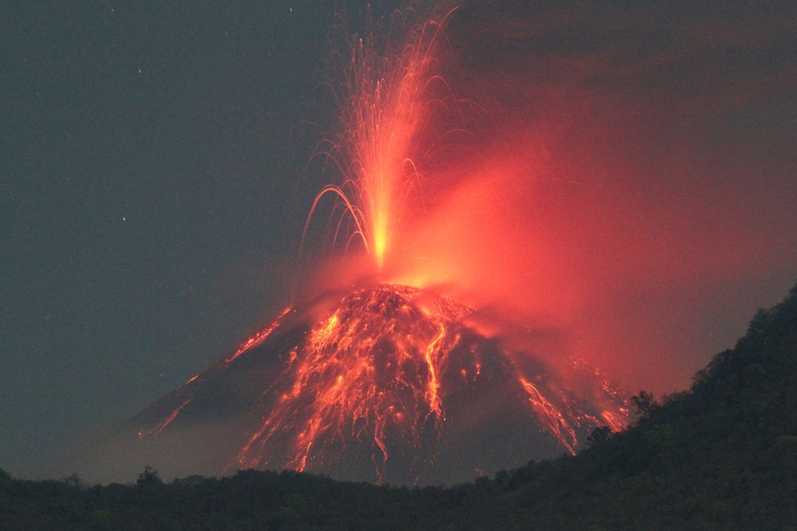 A volcano spits lava and gas, seen at night, with streams of lava flowing down its slopes.