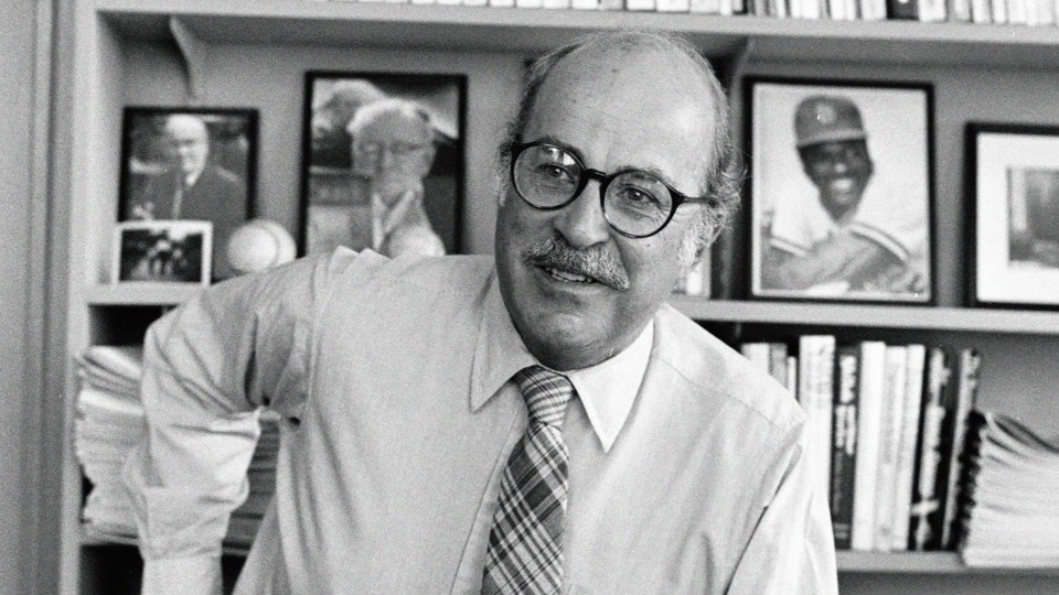 Black-and-white photo of the writer Roger Angell with baseball memorabilia and photos behind him on a bookshelf