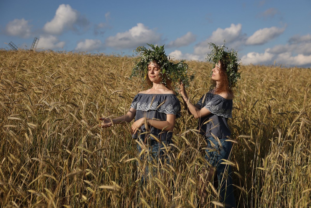 Two women wearing large wreaths of flowers stand in a wheat field.