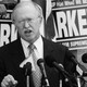 A black-and-white photo of a man speaking at a lectern with microphones. Behind him, people hold signs that read "Standing Up for What We Believe: Parker, Republican for Supreme Court."