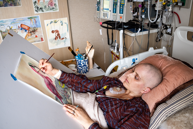 photo of woman with shaved head lying in hospital bed painting on easel with watercolors and hospital equipment on wall behind