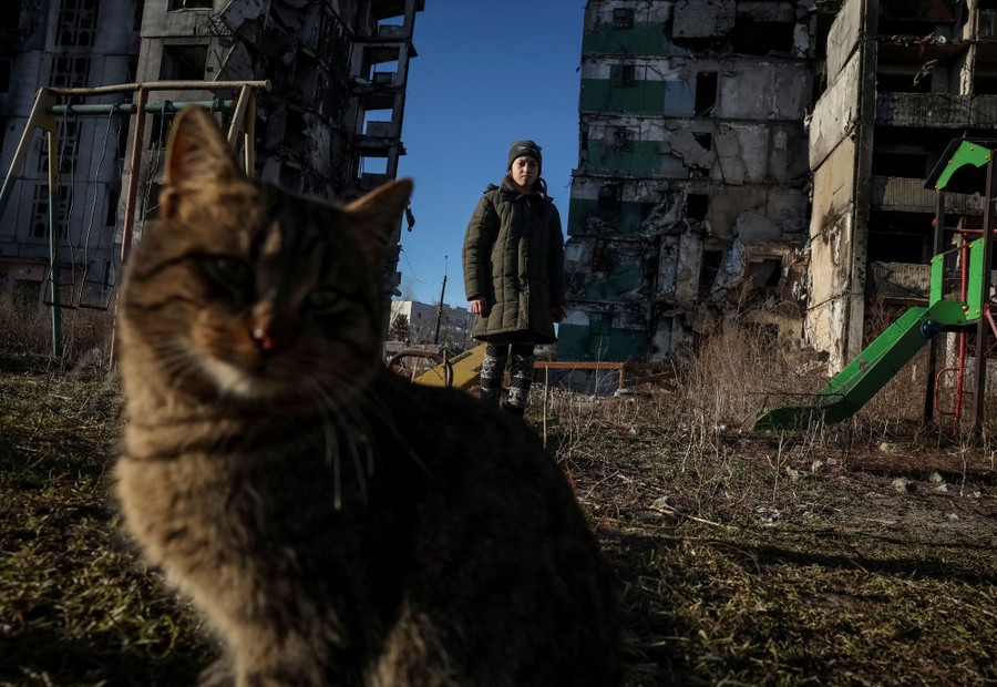 A girl stands near a cat beside a damaged building.