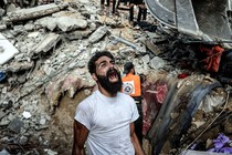 A man reacts, standing beside a huge pile of rubble following an air strike. Workers and onlookers stand nearby and atop the rubble.