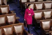 Elise Stefanik wears a pink suit and walks in front of empty seats in the House Chamber
