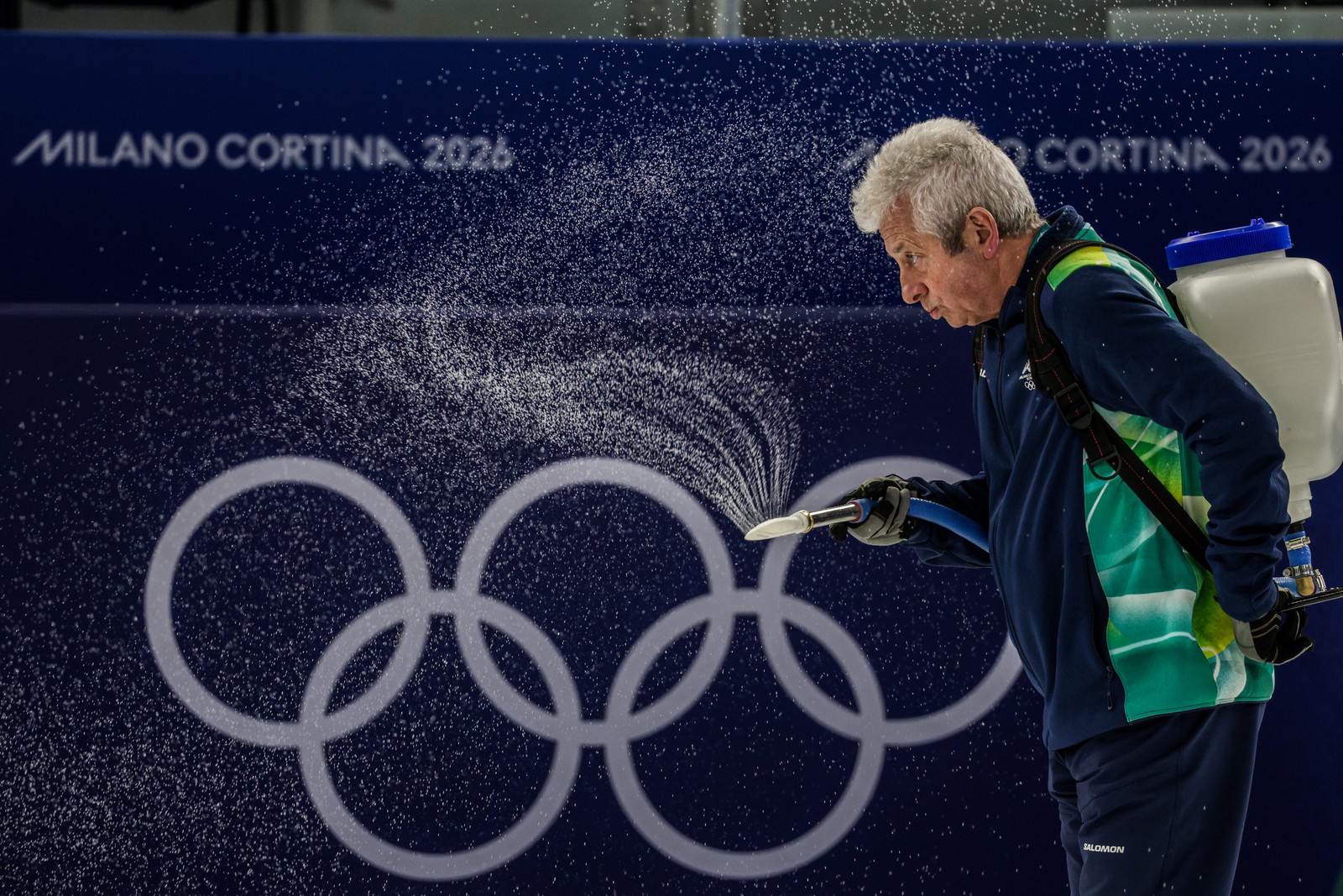 A worker wearing a tank on their back sprays water in an Olympic curling arena.