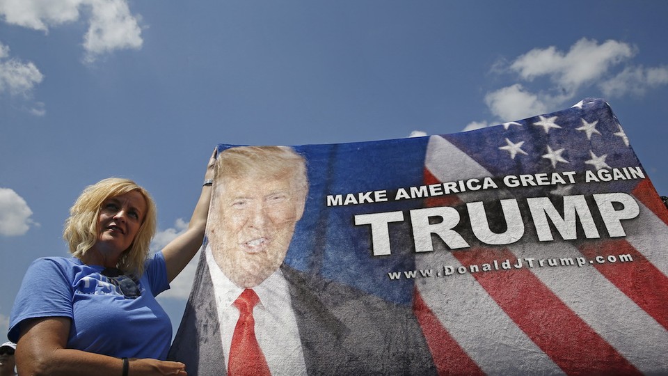 A white woman with short blond hair, wearing a blue T-shirt, stands outdoors holding up a Trump banner that reads "Make America Great Again."