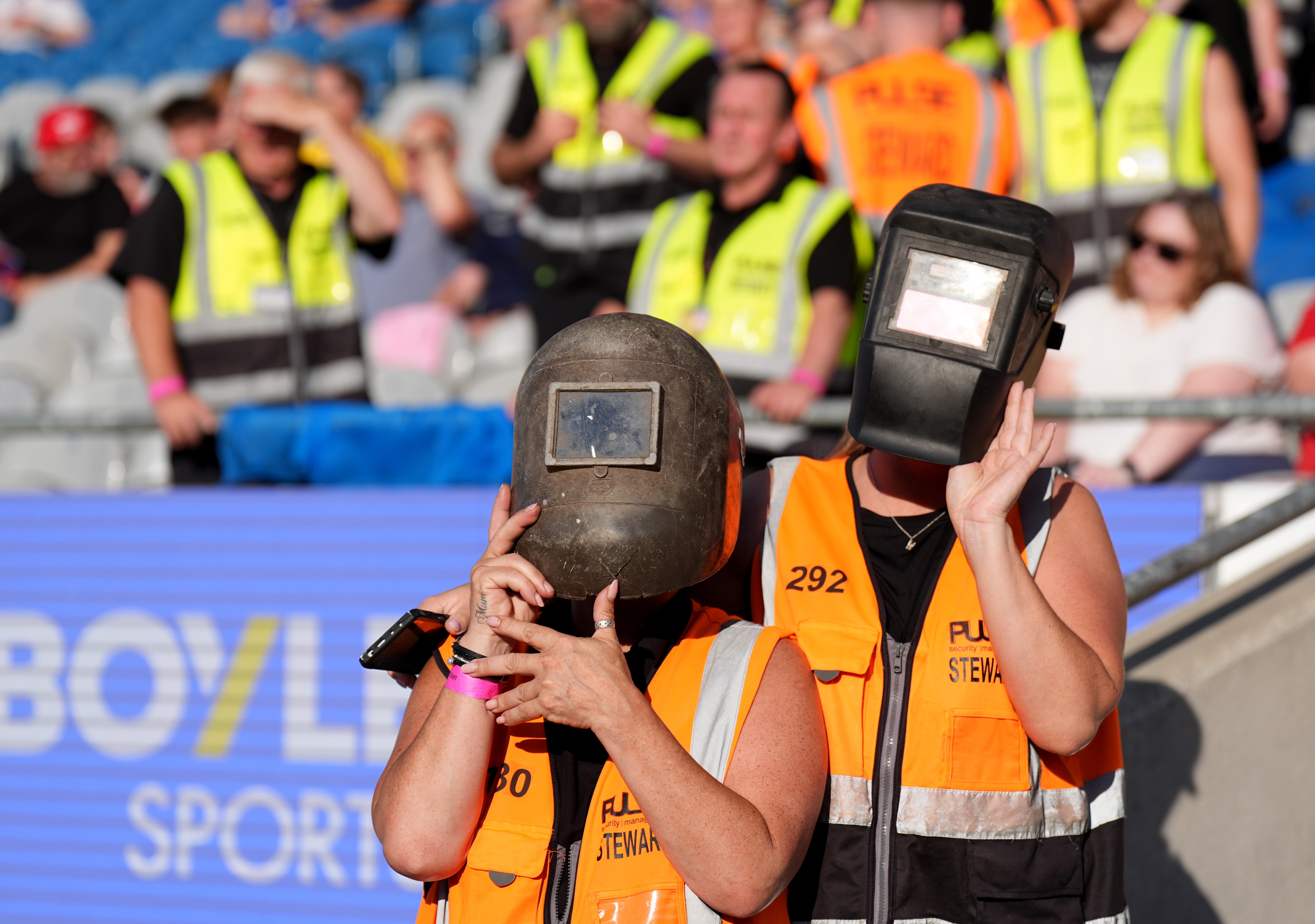 Two people wear welding helmets to watch a solar eclipse in a stadium.
