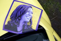 A purple poster of Kamala Harris rests on the hood of a yellow car