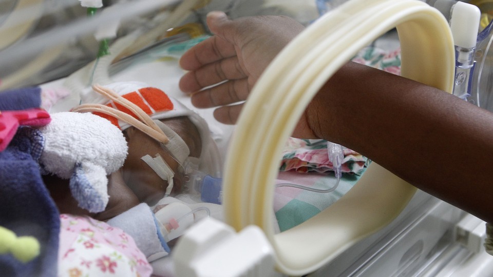 A hand reaches toward a premature infant in an incubator