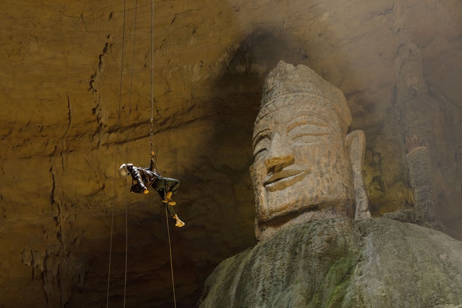 A person rappels into a cavern in front of a huge stone carving of a head.