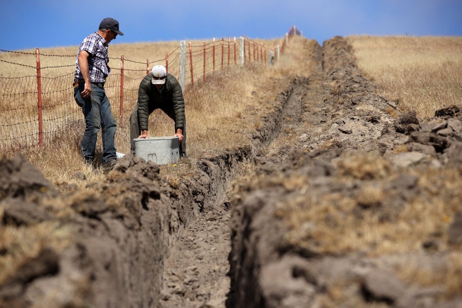Two people move a concrete box alongside a long trench dug in a pasture.