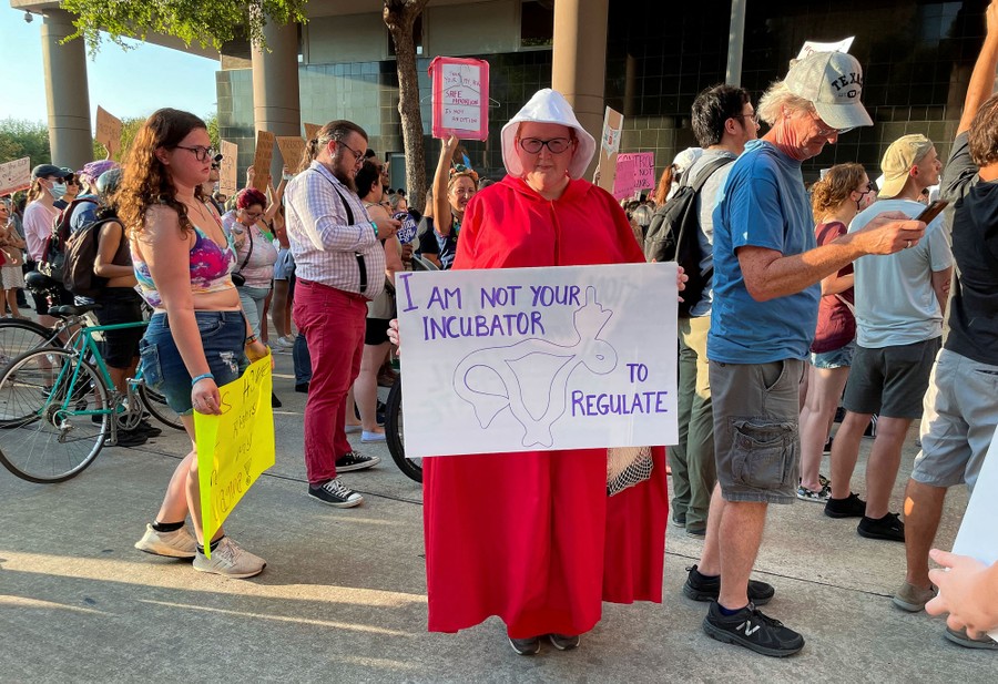A protester dressed in a red cloak and white cap holds a sign reading "I am not your incubator to regulate."
