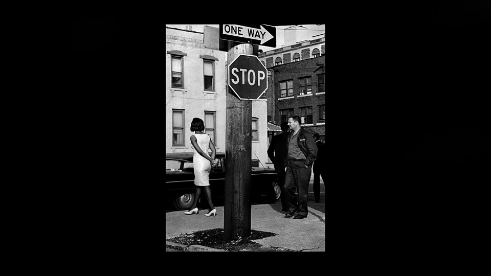 A black-and-white photo of a woman with her hands behind her back, facing away from the camera, and a man watching her