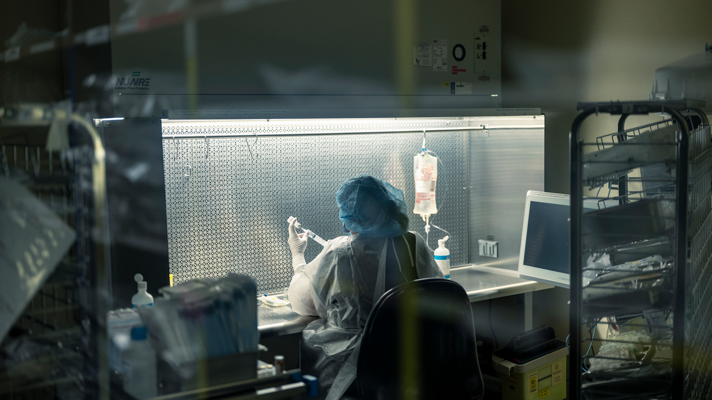 photo of person wearing protective medical gear and sitting at brightly lit desk in darkened room holding syringe