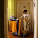 A hotel maid enters a room with a cart of cleaning supplies.