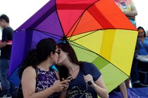 Two women kiss under a rainbow umbrella.