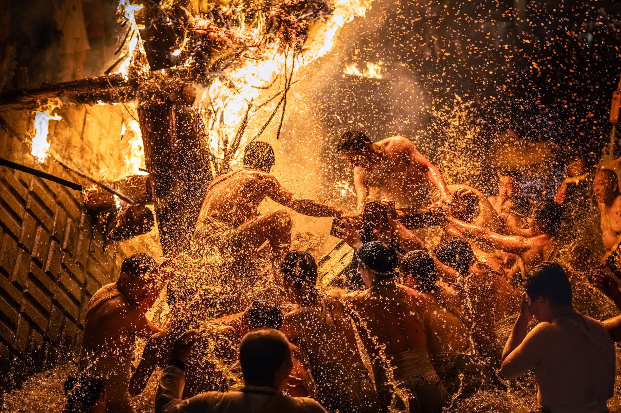 A chaotic scene during a festival, where men splash in a river beneath huge torches that send sparks flying.