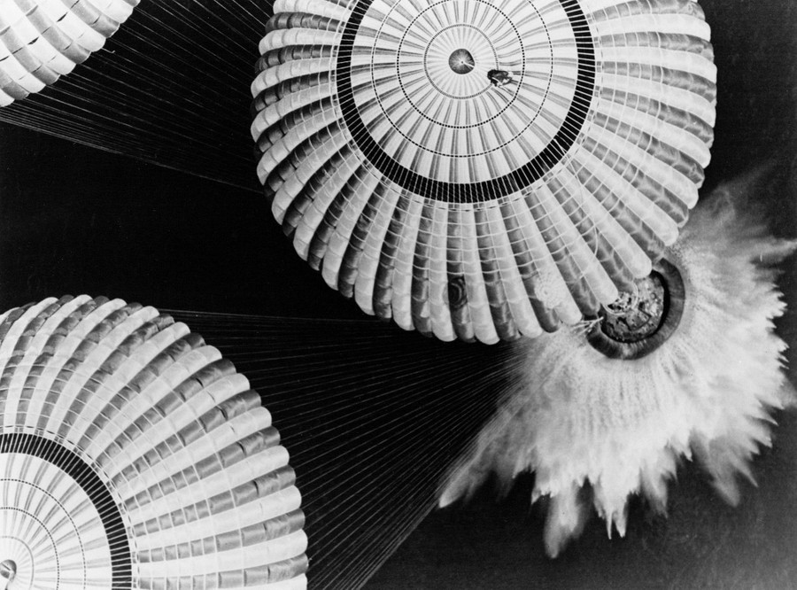 A view from a helicopter down on three large parachutes and the crew capsule as it splashes into the ocean