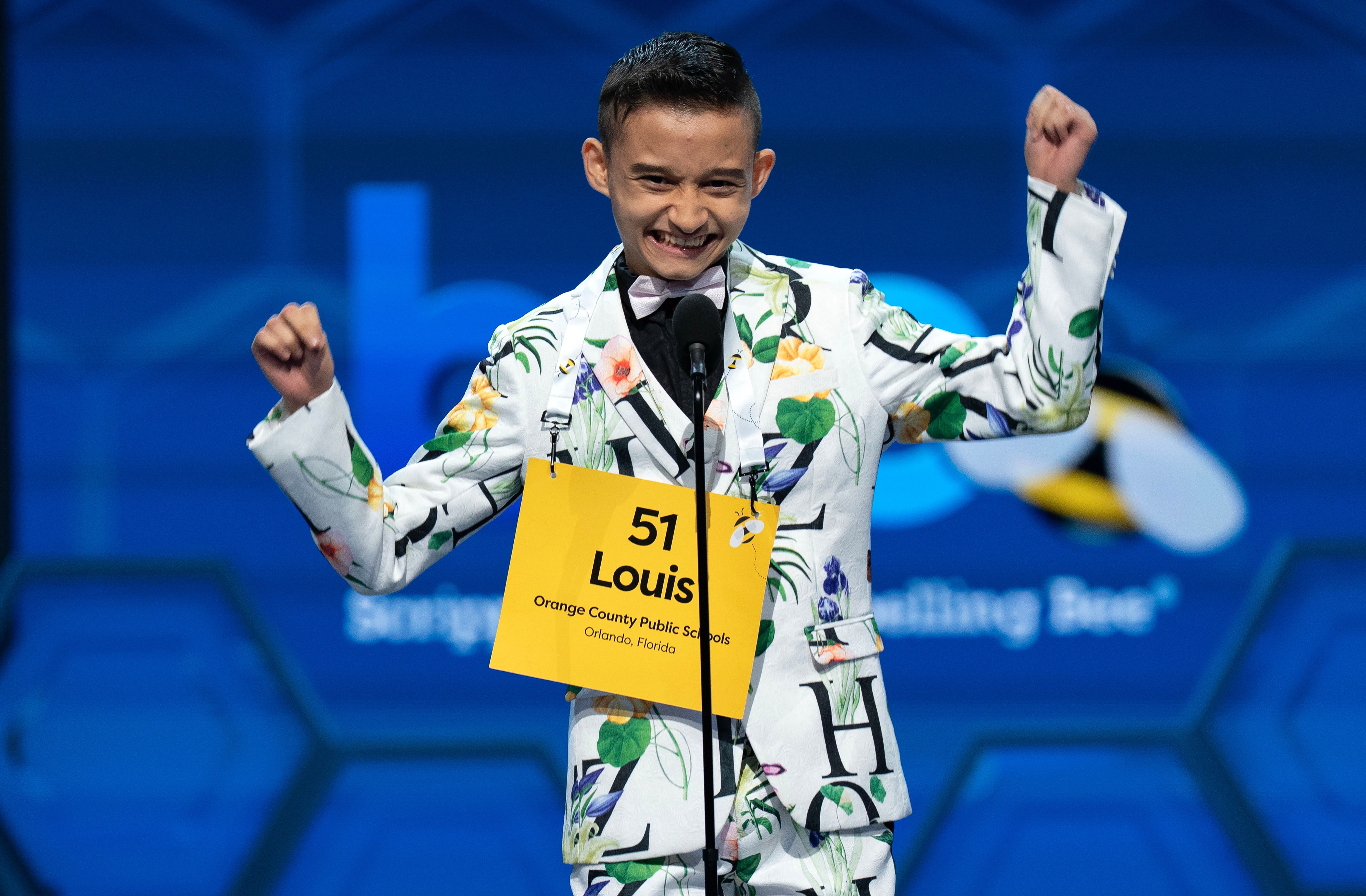 A young competitor at a spelling bee, wearing a suit made with a print fabric featuring letters and flowers, smiles and raises their arms to celebrate correctly spelling a word.