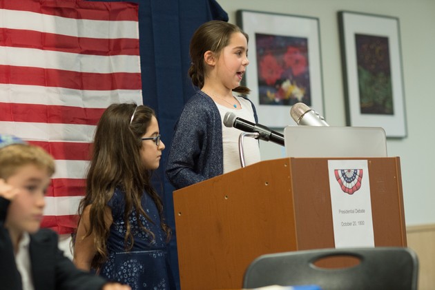 A third-grader speaks at a podium in front of an American flag.