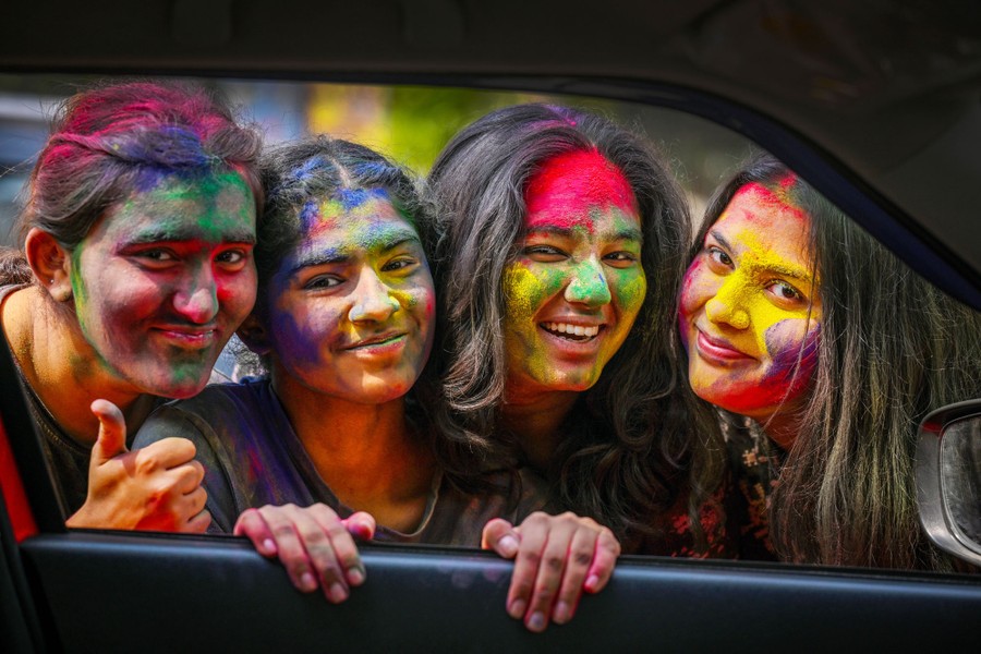 Four people with colored powder all over their faces smile and pose side by side, framed by the window of a car
