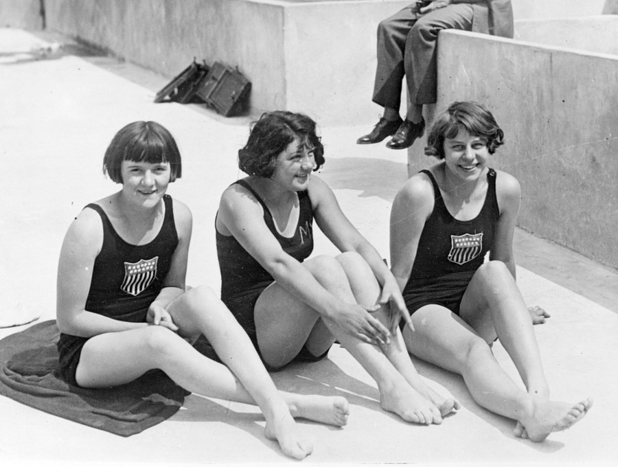 Three athletes in swimsuits sit together on concrete.