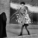 a woman in a headscarf walks in front of a mural showing Ruhollah Khomeini and toward a group of women in black chadors