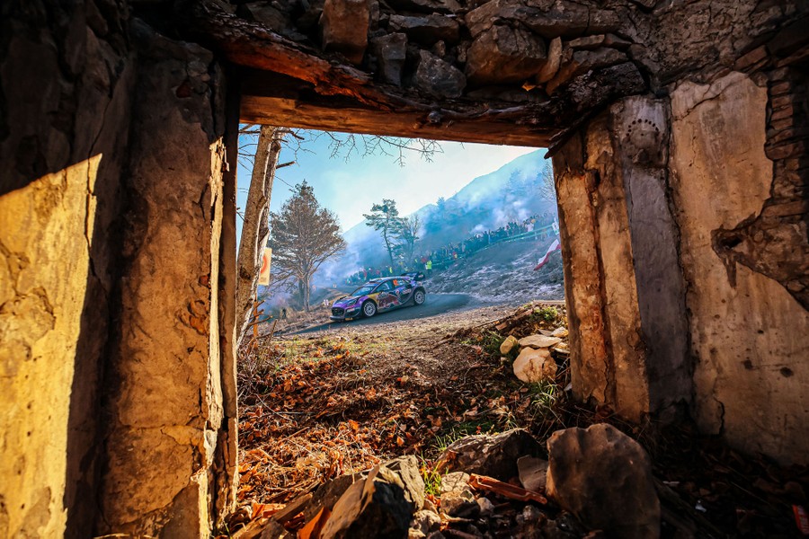 A view through an archway of a curved mountain road with a rally car racing on it.