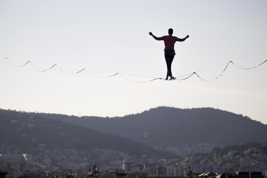 A tightrope walker walks on a rope high above a city square.