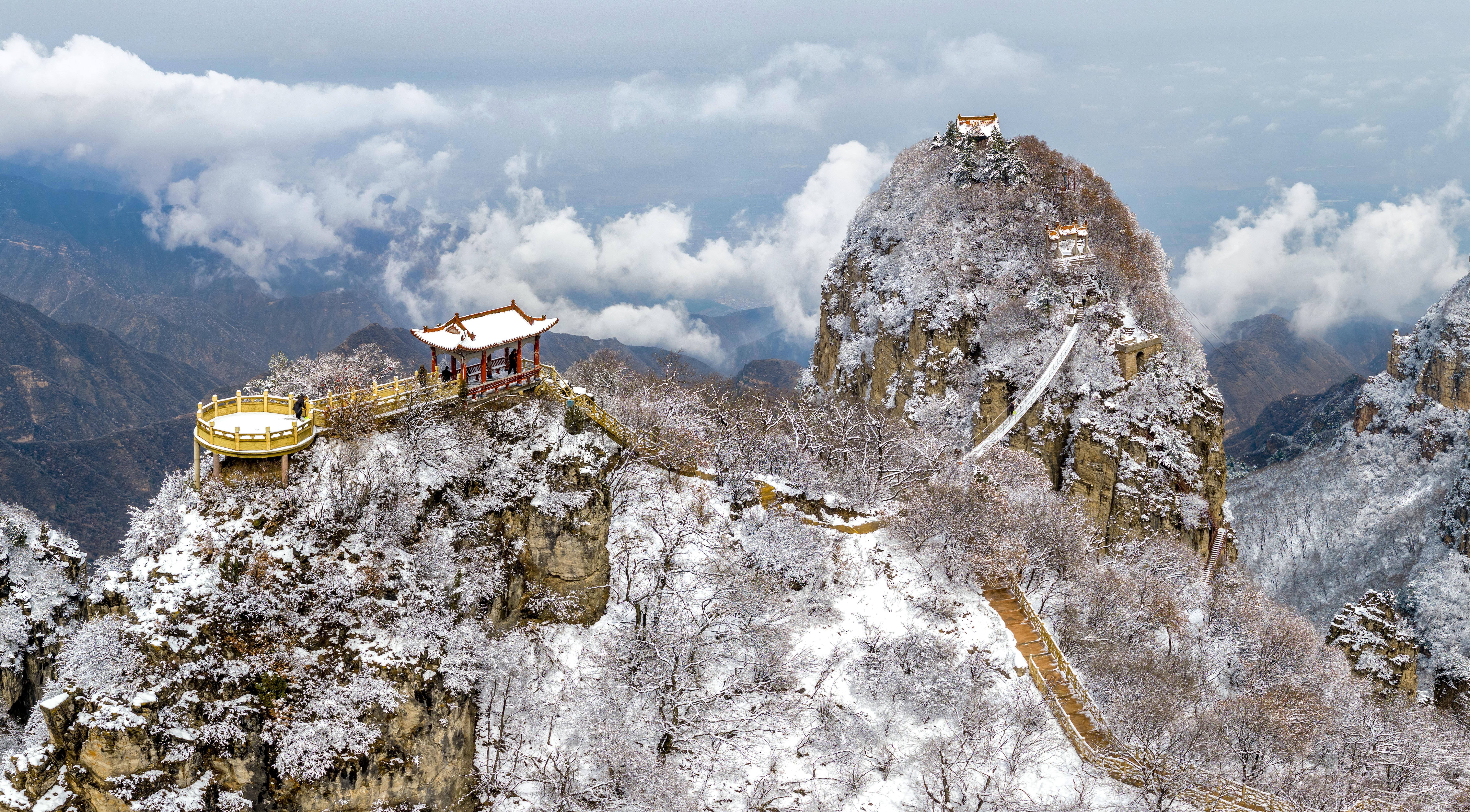 An aerial view of a steep set of snow-covered mountaintop rock formations, dotted with small temples and pathways.