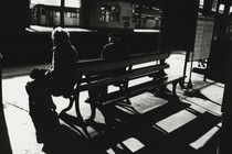 a black and white photo of two people sitting on a bench at a train station, from behind