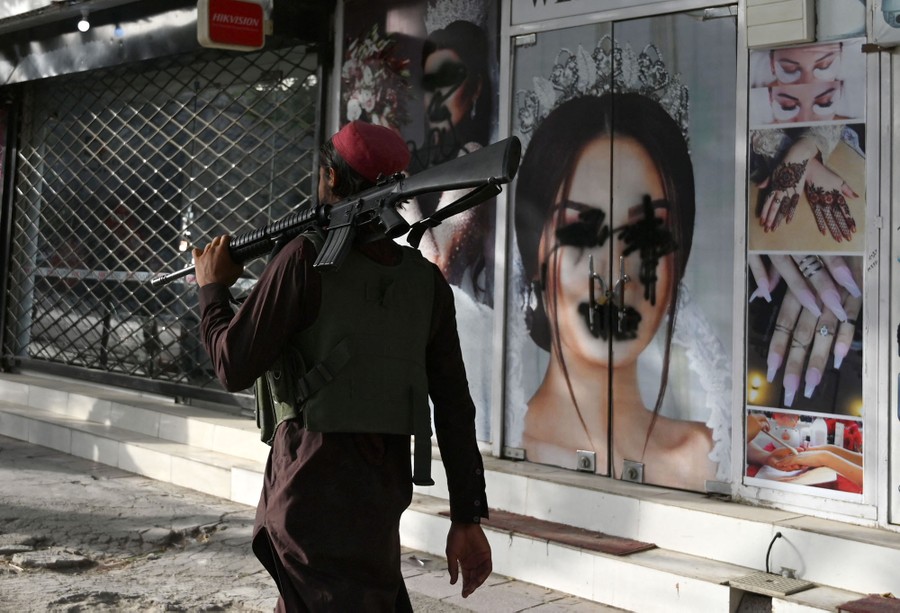A man with a rifle over his shoulder walks past a vandalized beauty parlor.
